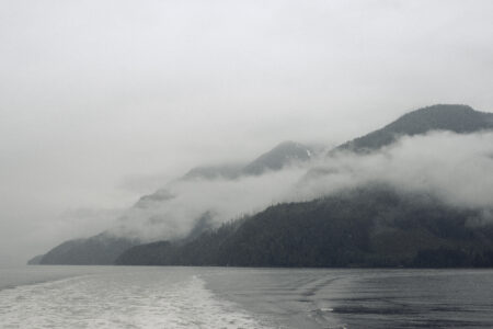 In Knights Inlet in the Great Bear Rainforest, the ocean is grey under an overcast sky. The water leaves a white trail behind a boat, and clouds hang low over green mountains