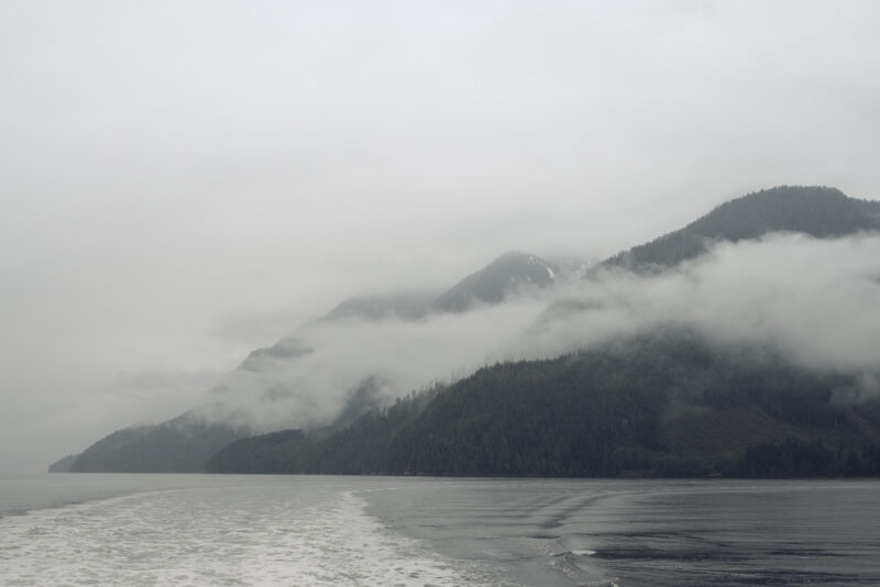 In Knights Inlet in the Great Bear Rainforest, the ocean is grey under an overcast sky. The water leaves a white trail behind a boat, and clouds hang low over green mountains