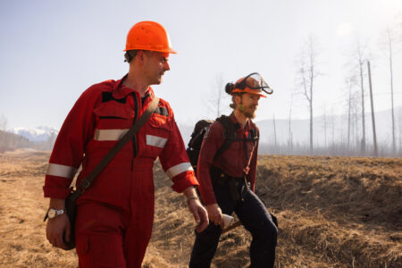 Matt Simmons walks with another person, both wearing hard hats and fire protection gear, through a smoky landscape.