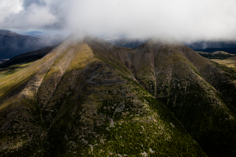 An aerial view of the Klinse-za (Twins Sisters) Mountains. The majestic mountains are cast in sunlight and shadow, green dotted with yellow at the bottom and brown near the top, with the very top peaks obscured in clouds.