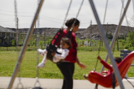 A mother pushes two small children on a swing set, with mounds of landfill waste in the distance