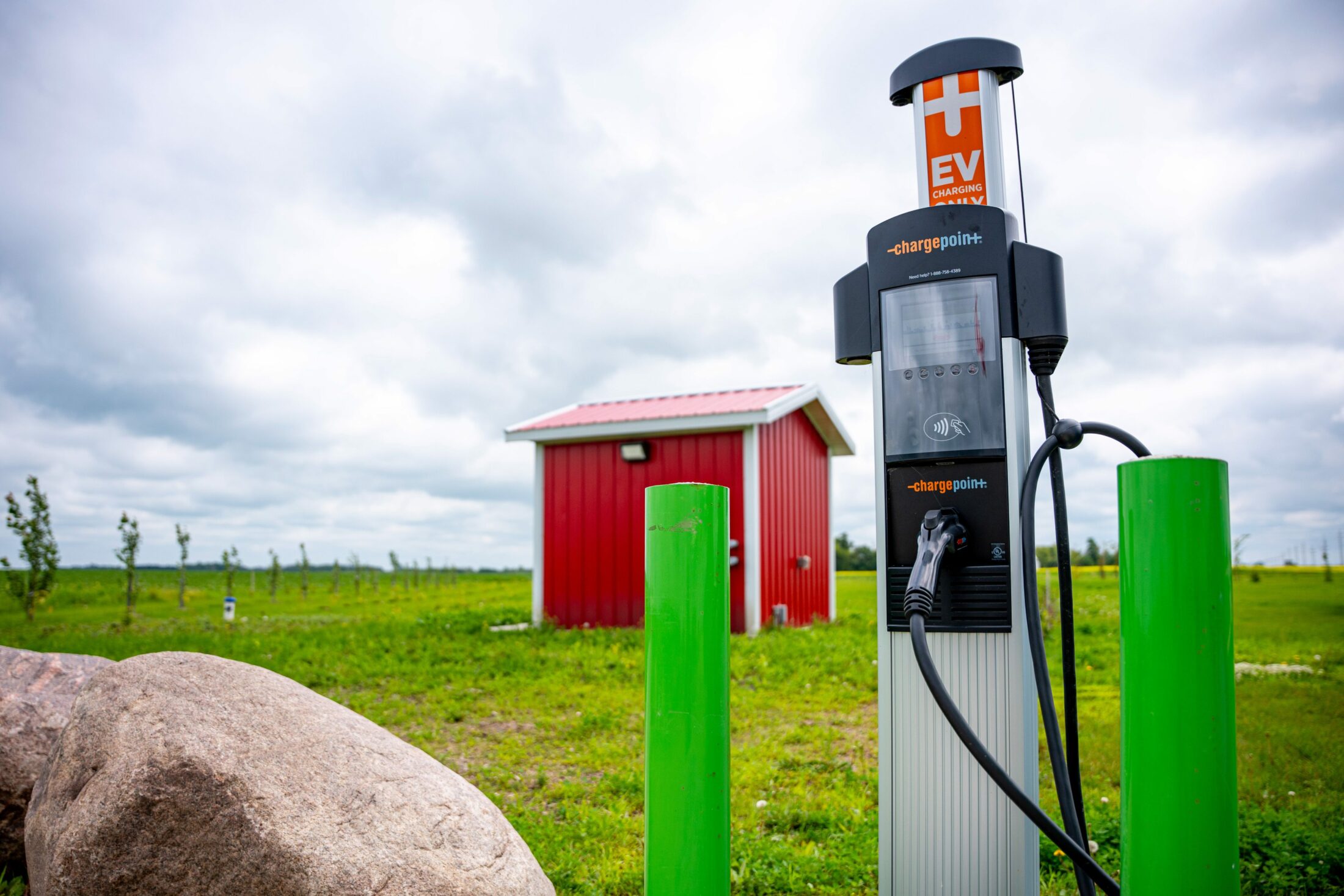 A charge point electric vehicle charger flanked by green posts in front of a small red building in rural Manitoba