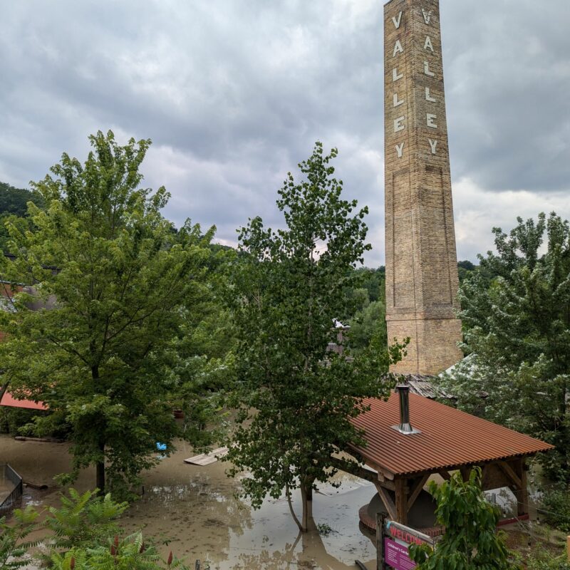 The old brick smoke stack at the Brick Works site towers over trees and a rooftop with floodwater pooled below