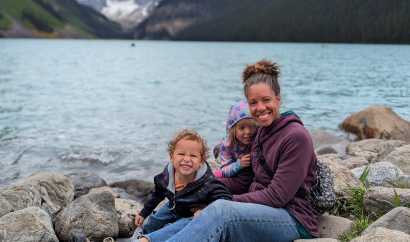 Brittany Hopkins with her children in Banff, AB, sitting on a rocky shore of a lake with mountains behind.