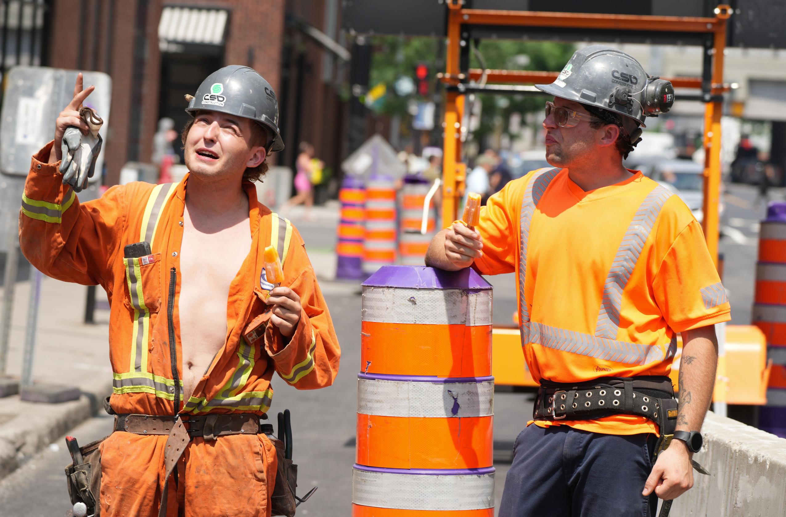 Heat wave: two construction workers, one with his coveralls unzipped to his waist, eat popsicles and gesticulate