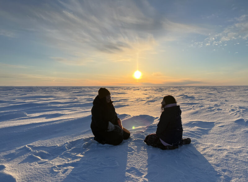 Two youth sit on the sea ice in front of a setting sun in Tuktoyaktuk, NT