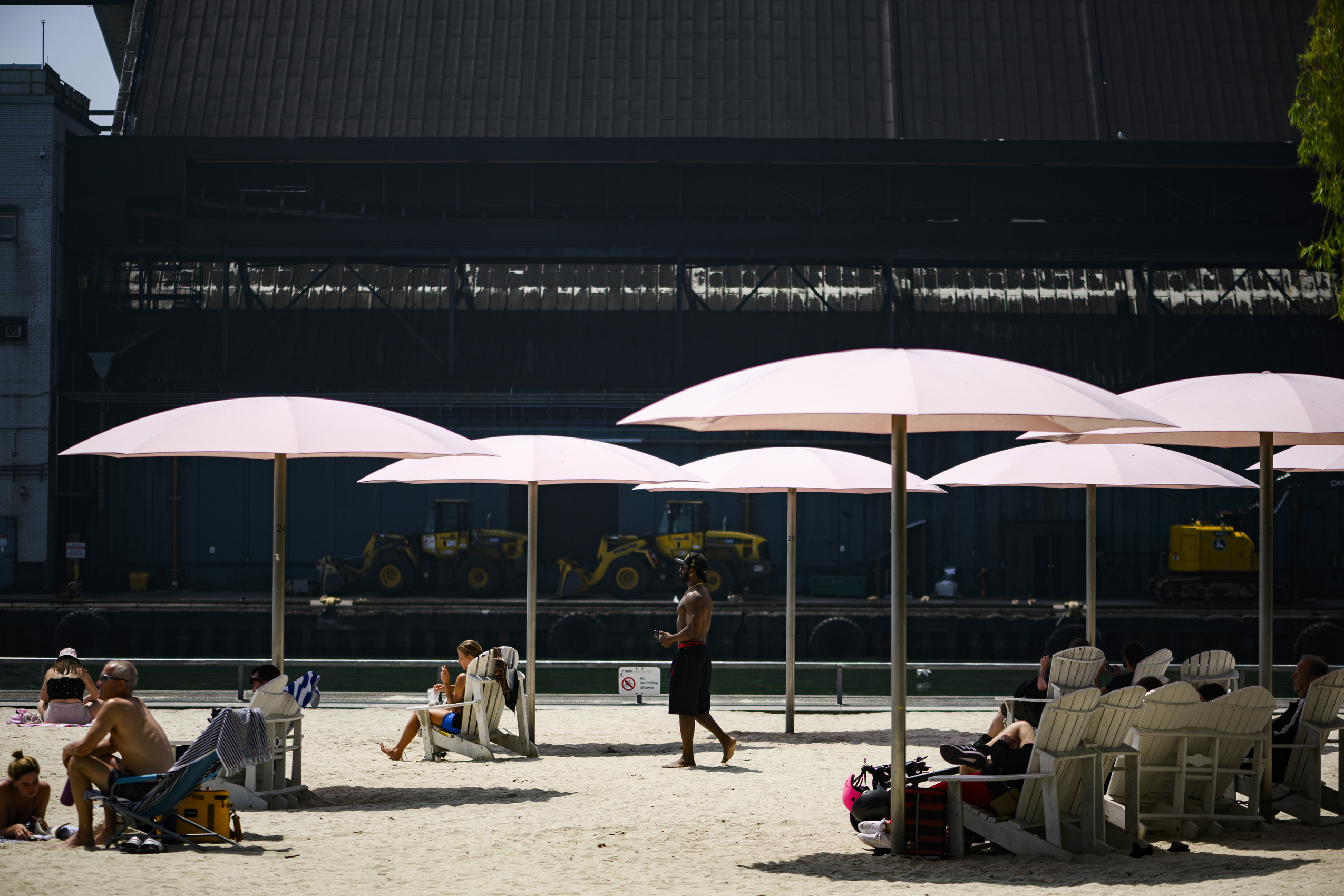 Heat wave: people lounge in beach chairs under pink umbrellas in the sand with a factory in the background