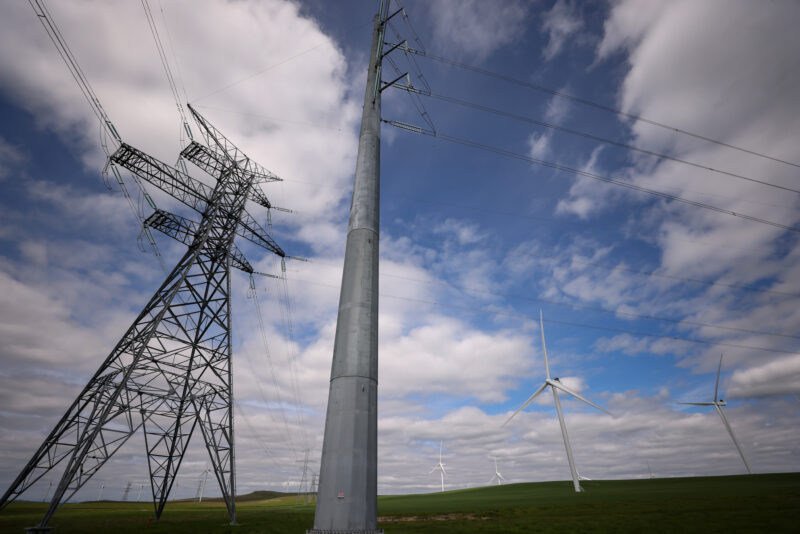 Transmission lines rise up from the ground, with wind turbines in the background.