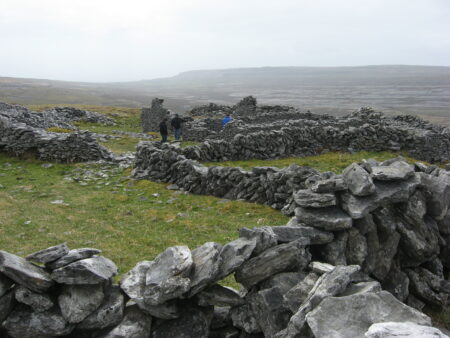 Flat grey stones are stacked in long lines delineating homes long ago abandoned, on a grassy field under foggy skies