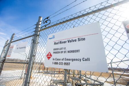 A chain-link fence with barbed wire in front of an industrial site. A sign on the fence reads "Red River Valve Site" and includes a number to call in case of emergency