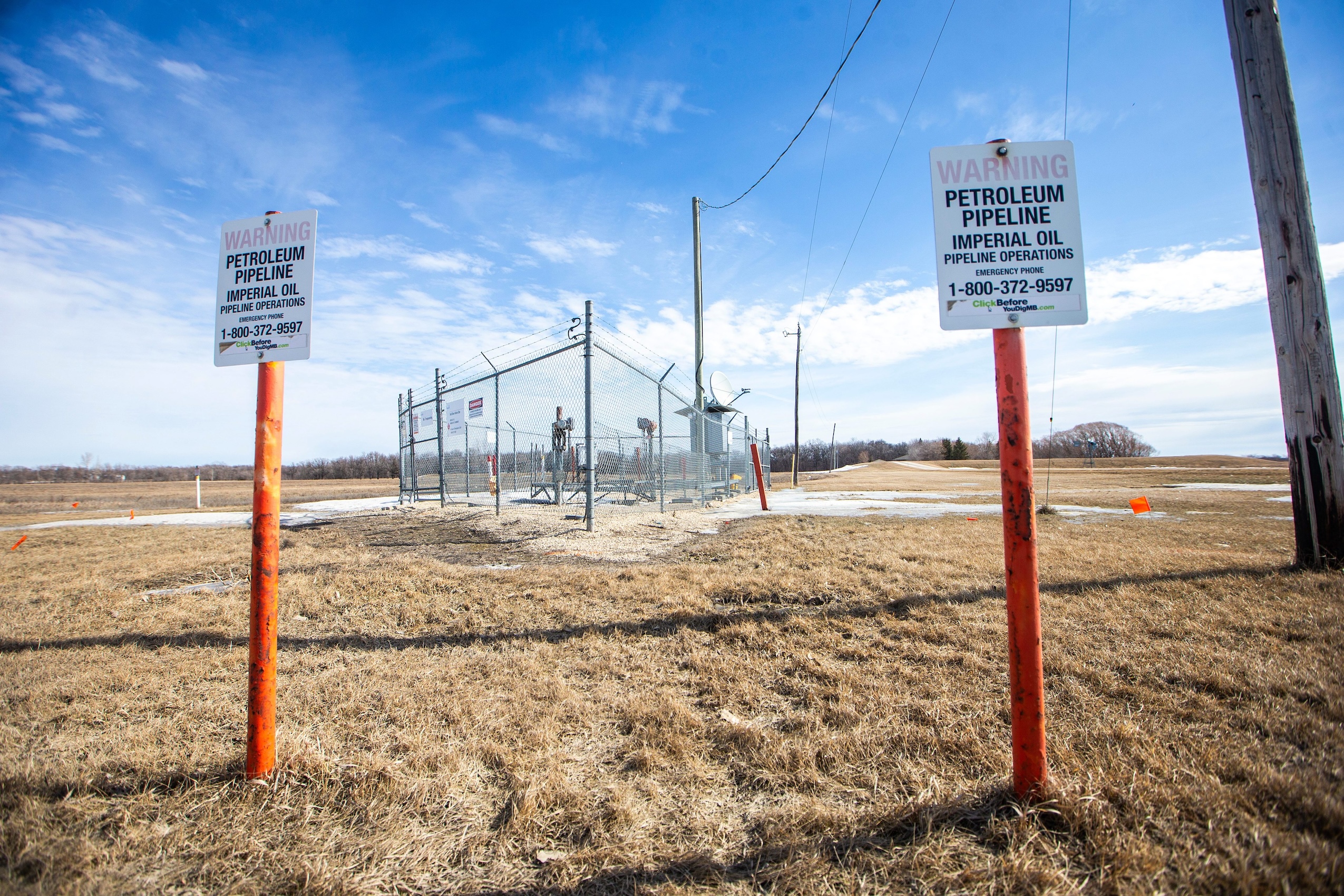 Two signposts warn the presence of a petroleum pipeline. Behind, above-ground pipeline infrastructure is surrounded by chain-link fence and barbed wire.