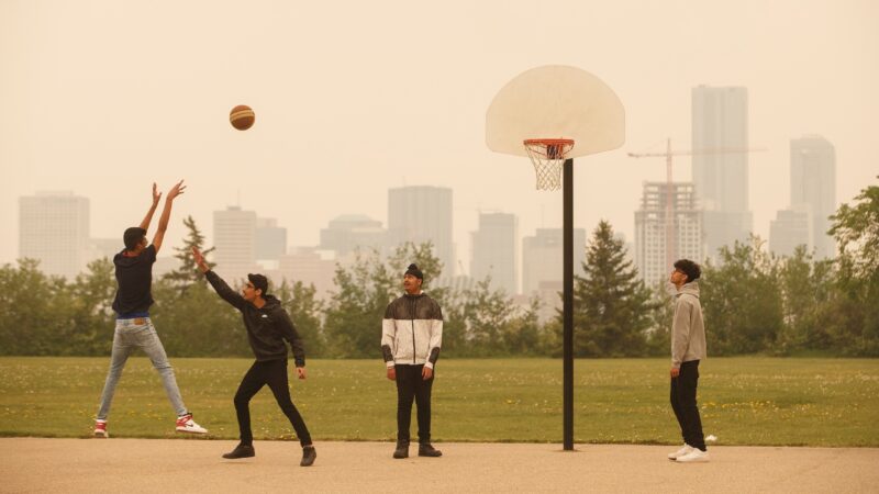 Four young people play basketball as one shoots towards the net. Behind, an urban skyline is shrouded in wildfire smoke.