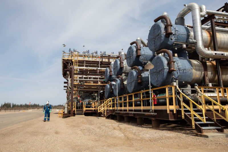 The Cenovus Christina Lake oilsands facility is seen on a dirt field with a man in a blue jumpsuit in the background.