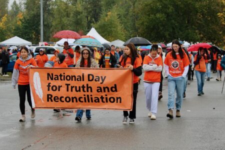 Dozens of people in orange shirts walk down a street. They are led by an orange banner that reads, "National Day for Truth and Reconciliation."