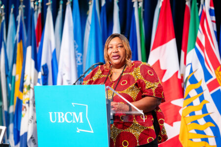 Union of BC Municipalities president Trish Mandewo stands at a podium in front of a bank of flags, including the Canadian flag