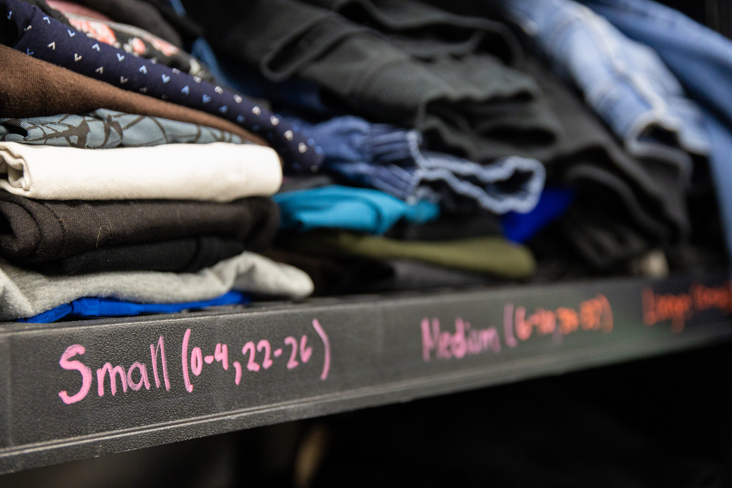 A close up of a storage shelf piled with various clothing items arranged by size in the back room of a Winnipeg thrift store. A label on the shelf reads “Small (0-4, 22-26)” in pink lettering