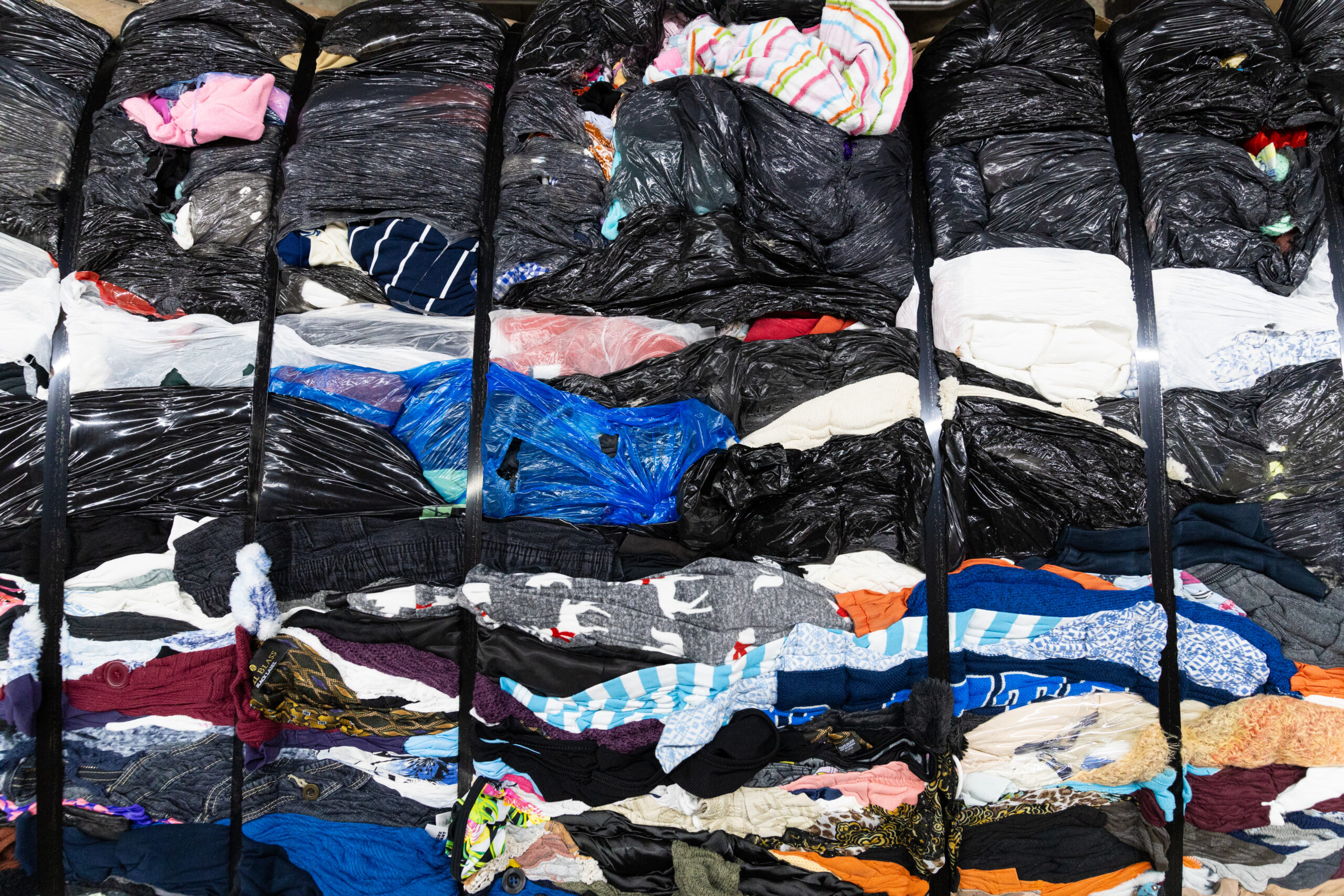 Multi-coloured clothing items and black plastic garbage bags compressed into a clothing bale and tied together with black straps