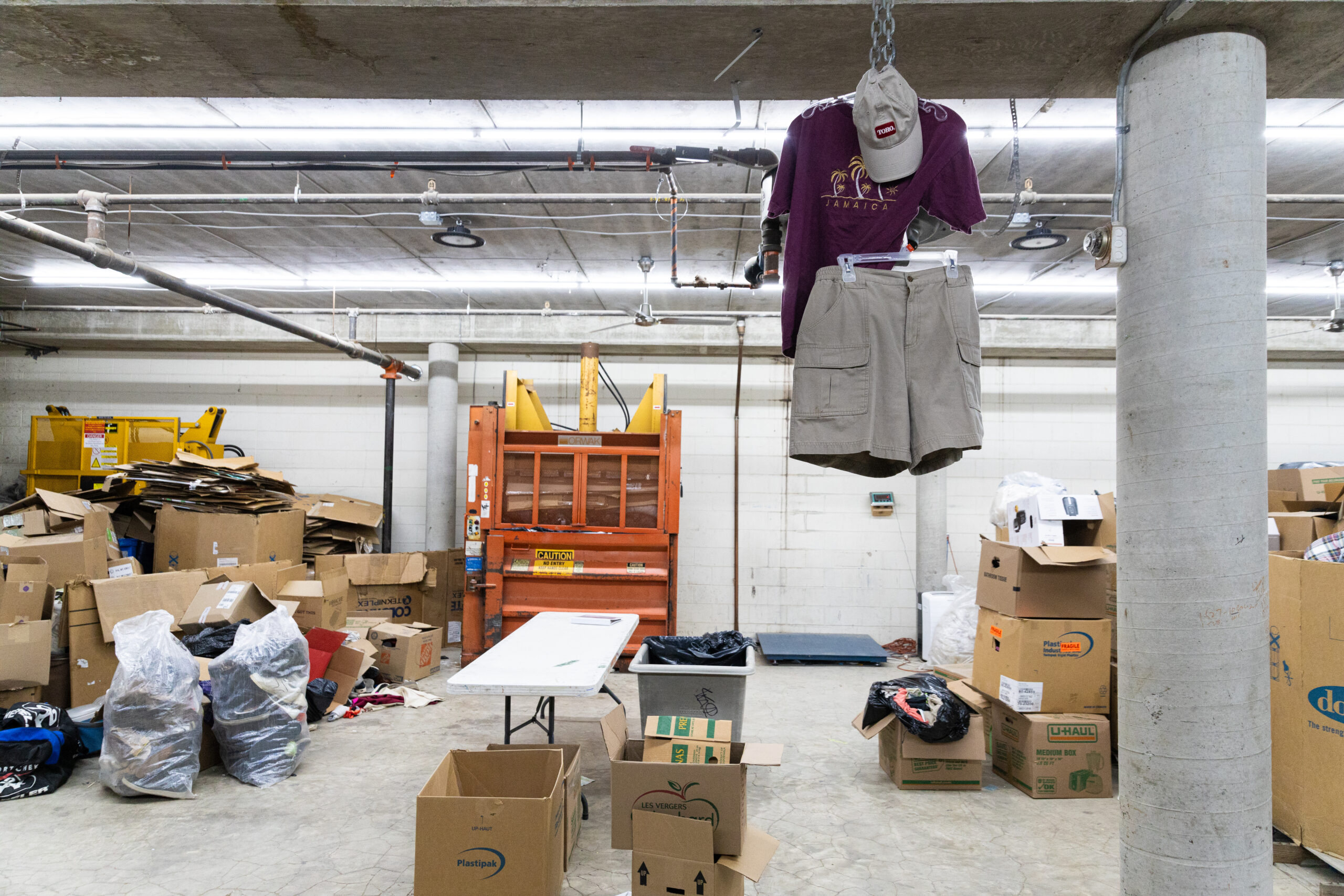 A beige cap, beige cargo shorts and purple graphic tee hang from a chain next to a large concrete pillar in the foreground. In the background, a large orange baling machine is flanked by cardboard boxes and plastic bags of clothing in a Winnipeg thrift warehouse
