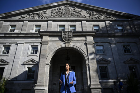 Woman in blue suit stands in profile of dark shadow in doorway of large stone building