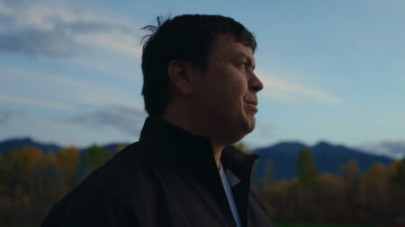 A portrait of Jacob Beaton at Tea Creek, facing the soft light of sunset or sunrise. He wears a black sweater and looks into the distance, with mountains and a blue sky with wispy white clouds in the background. Jacob Beaton is pursuing Indigenous food soveeignty at his farm, Tea Creek.