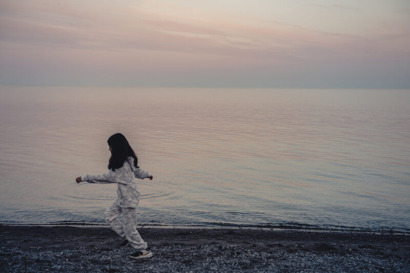 A child skips along the shore of Lake Ontario with a pink sunset on the horizon behind her