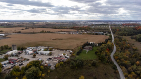 Highway 413: an aerial view of a road winding past a forest, a farm and a parking lot, headed towards a suburb on the horizon