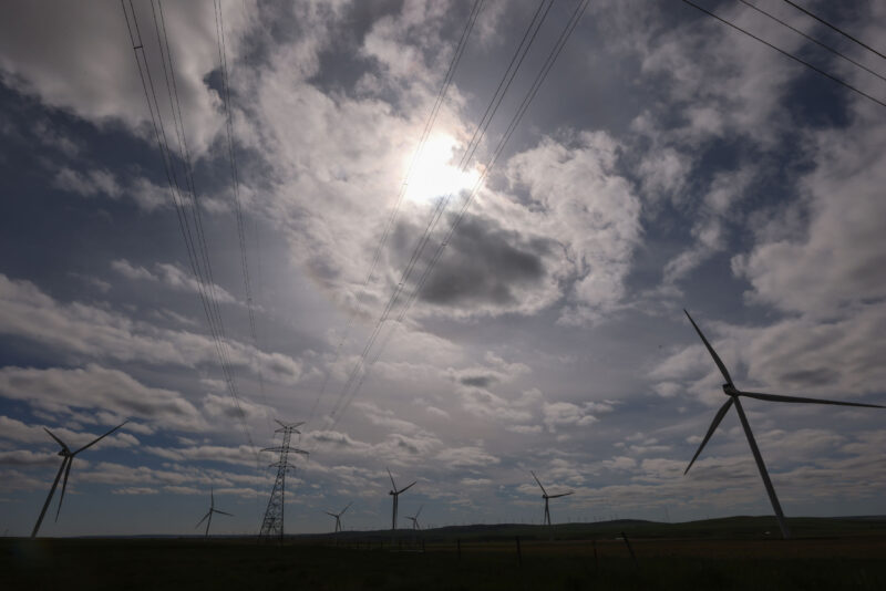 Looking up at wind turbines and large electricity transmissions lines set against a big sky with a few clouds obscuring the sun