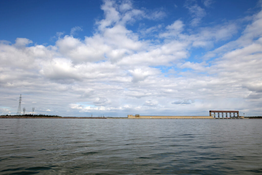 The Limestone hydroelectric dam blocks the path of the Nelson River in northern Manitoba. The spillway gates jut upward on the right side while the low beige generating station stretches across the river to its left