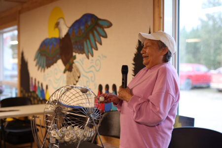 A woman holding a microphone laughs while calling bingo in front of a paiting of an eagle on the wall