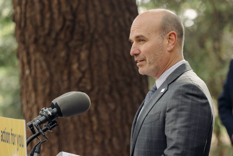 Nathan Cullen, speaking at a podium outdoors, with a tree in the background