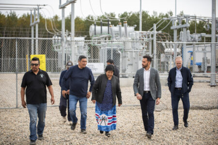 A group of five men and one woman walk away from a substation. The woman in the centre of the group, wearing a blue skirt with the logo of a transmission line company on it.