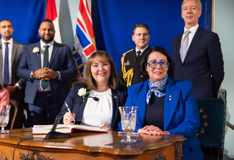 North Coast-Haida Gwaii MLA Tamara Davidson sits at a table with Janet Austin, B.C.'s Lieutenant Governor. Davidson, wearing a white, high necked shirt and navy blazer with a white rose on the lapel, is signing the book as Minister of Environment and Parks