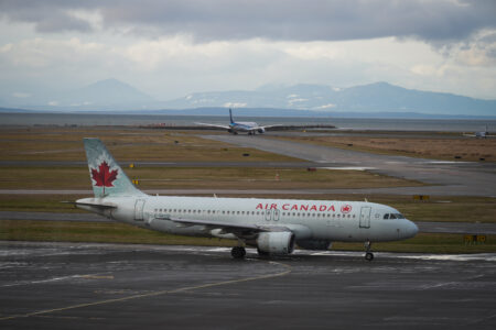 An Air Canada plane sits on a runway at Vancouver International airport. In the background, another plane is taxing down a runway that faces the water