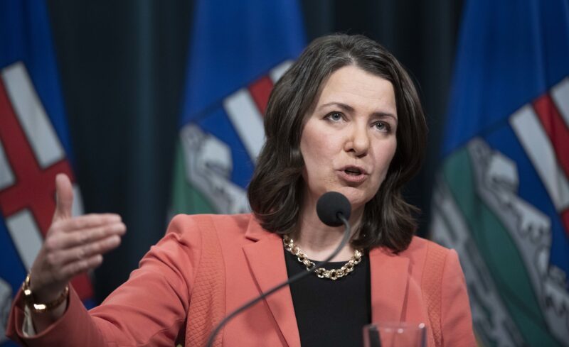 Alberta Premier Danielle Smith in front of a microphone, gesturing with her hand with an Alberta flag in the background.