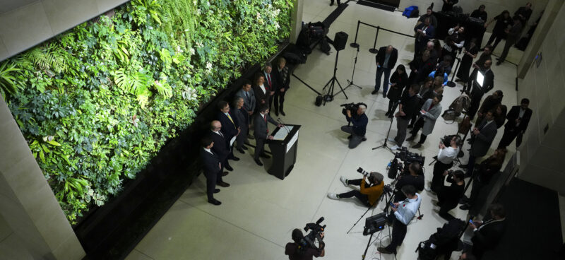 Green wall with people standing in front of it giving a press conference.