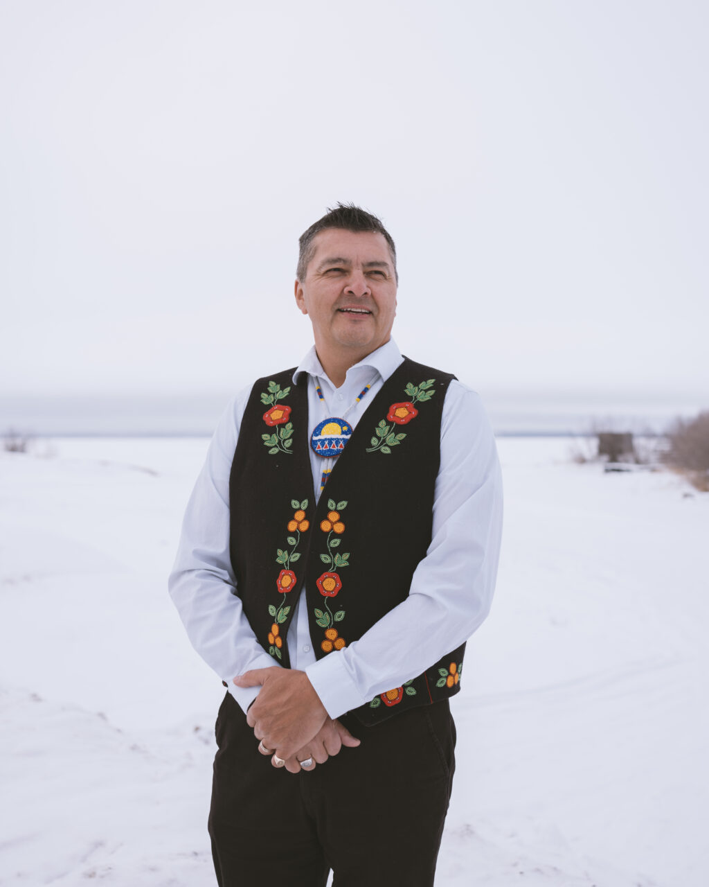 Jackson Lafferty stands in the centre of the image, wearing a beaded vest and medallion, with a snowy plain behind him.