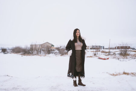 A woman in a fur-lined parka stands in a snowy landscape with a few houses in the distance