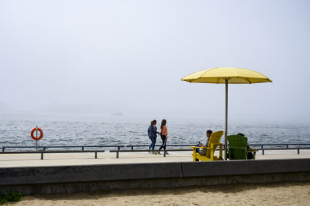 People sit in two chairs under a yellow umbrella on Lake Ontario in Toronto, with two people walking on the boardwalk in front of them, and the lake beyond.