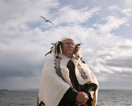 Hereditary Chief Eric Pelkey wears wool regalia and looks intently into the camera. He wears white wool regalia with brown accents. The sunlight comes from the fight and illuminates the soft wool, his right cheek and his white hair. The ocean in the background and the cloudy blue sky are awash with light.
