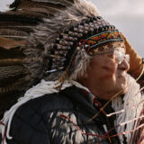 Hereditary Chief Paul Sam Sr. looks into the distance to the right, and sun bathes his face from that direction. He has red ocher paint (tumulh) on his face. The sunlight is soft on his face and reflects in his glasses. He wears traditional regalia and holds one hand to the side of his face, resting on his feather headdress