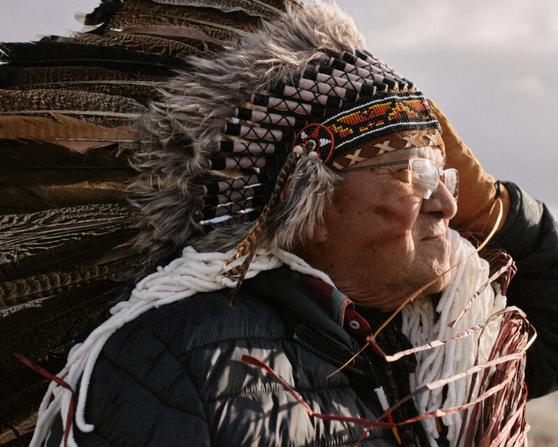 Hereditary Chief Paul Sam Sr. looks into the distance to the right, and sun bathes his face from that direction. He has red ocher paint (tumulh) on his face. The sunlight is soft on his face and reflects in his glasses. He wears traditional regalia and holds one hand to the side of his face, resting on his feather headdress