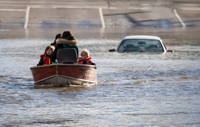 In the Abbotsford 2021 flood, a woman and her children are in a small motorboat on a water-filled road, being rescued by a volunteer. The smaller child has a soother in their mouth. A half-submerged car is visible behind them.