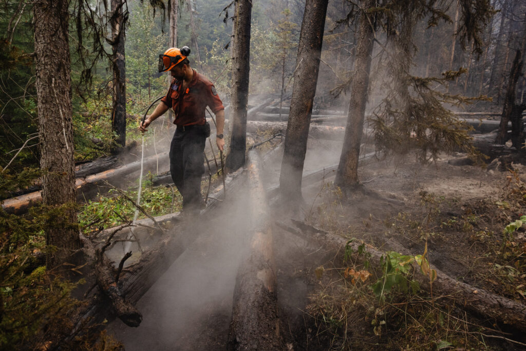 BC Wildfire Service firefighter standing on a log, spraying the ground, smoke all around