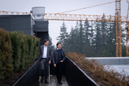 Man in suit walks with man in fall jacket against backdrop of construction crane and industrial building.