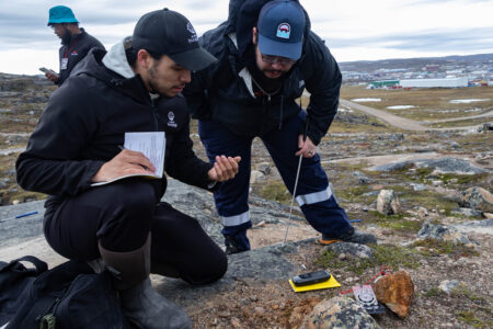 Two men crouch down looking at a rocky outcrop, with a notebook, compass and GIS in front of them