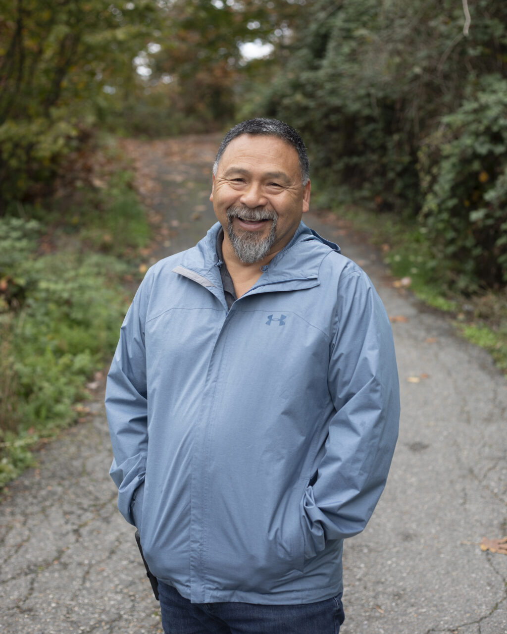 Murray Ned, wearing a blue coat, smiles while standing on a road with trees behind him.
