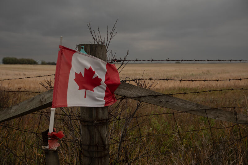 A small Canadian flag on attached to a barbed-wire fence with prairies in the background.