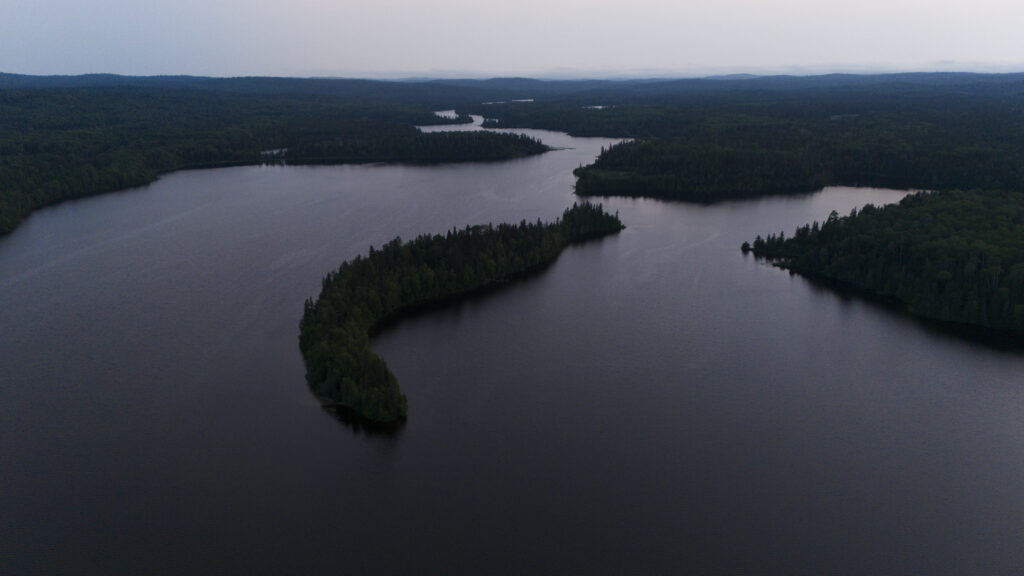 An aerial view of a lake with a forested island in the centre. Rolling hills blanketed with trees extend towards the horizon in the background