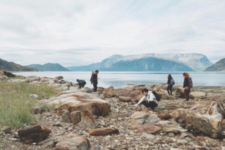 Several people meander on a scenic rocky beach with low tundra mountains in the distance. Some crouch down to look at rocks