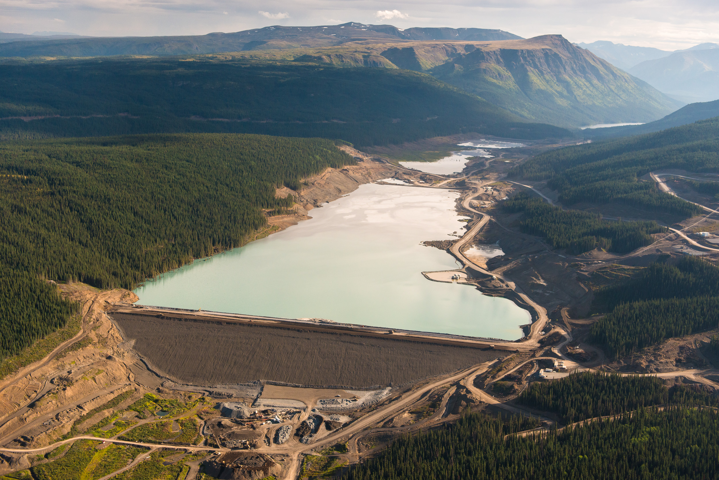 An aerial view of the tailings pond at the Red Chris Mine. The wall of the massive pond dominates the view and the large industrial vehicles driving along its top look like ants